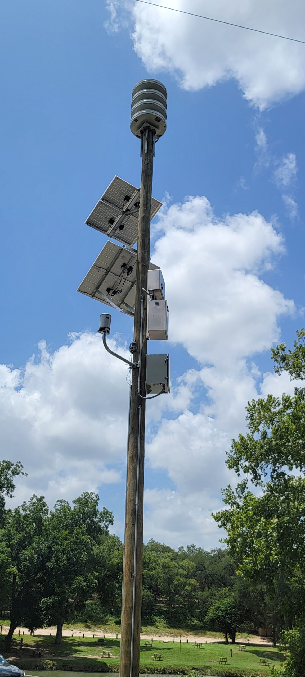 A complete AWARE warning installation on a utility pole beside a park and river, with an omnidirectional siren at the top, four solar panels, a camera arm, and equipment cabinets