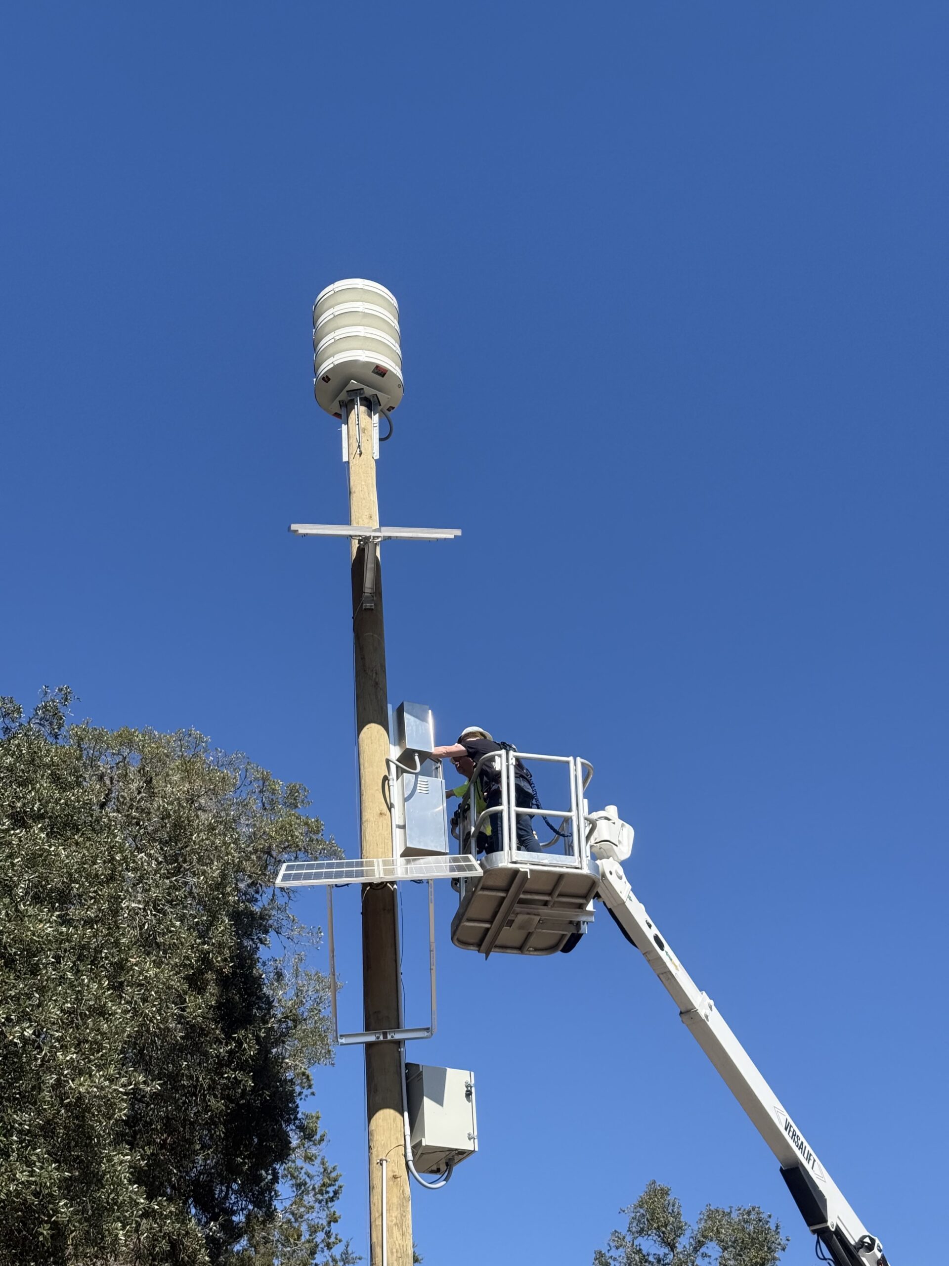 A technician working from a bucket truck to install an outdoor warning siren array on a pole, with solar panels and equipment cabinets mounted below