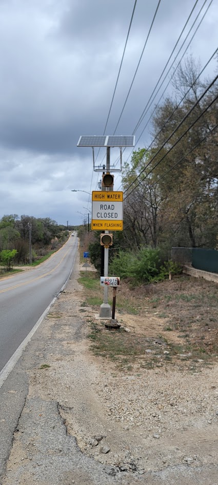 A solar-powered low-water crossing warning system beside a rural road, with a High Water Road Closed When Flashing sign and amber warning beacons